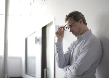 Man with glasses leans against a wall, looking stressed after financial loss.