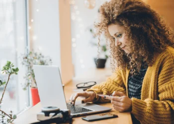 Woman in yellow sweater shops online on a laptop. Emotional spending adds up.