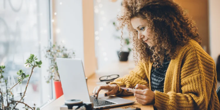 Woman in yellow sweater shops online on a laptop. Emotional spending adds up.