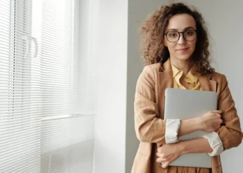 A woman stands beside a window and holds a laptop in her arms. SMART Goals can help you reach the finish line.