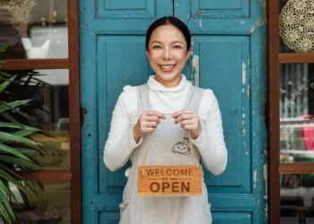 A woman stands in front of a blue door with a sign that says, "Welcome we are open." Small business finances are important to understand.