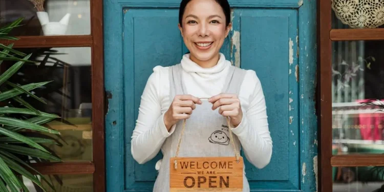 A woman stands in front of a blue door with a sign that says, "Welcome we are open." Small business finances are important to understand.