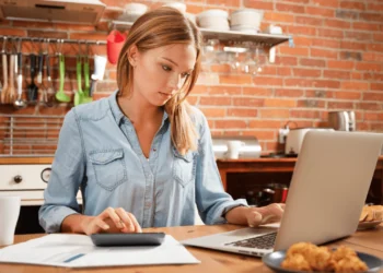 A young woman sits at a table, looking at a laptop screen at different types of budgets.