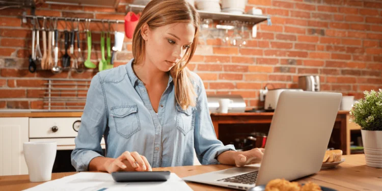 A young woman sits at a table, looking at a laptop screen at different types of budgets.