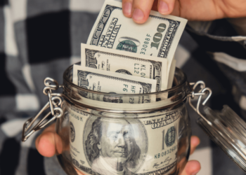 Someone holds a glass jar full of American cash and adds $100 to it. Personal finance tips to help you finish 2024 on a strong note.