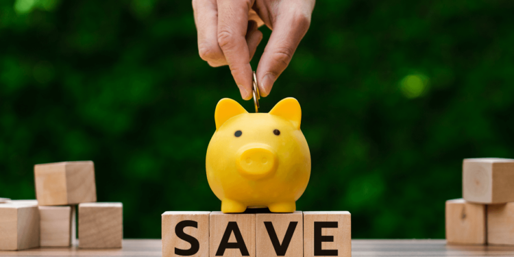 A hand drops a coin into a yellow piggy bank that's standing on wooden blocks that spell out SAVE. A high yield savings account can help you save faster.
