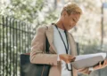 A young woman in a camel coat stands outside a fence looking at a folder. Financial success stories to inspire you.