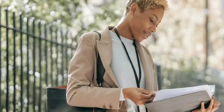 A young woman in a camel coat stands outside a fence looking at a folder. Financial success stories to inspire you.