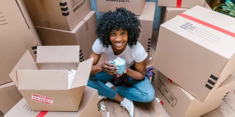 A young woman in a t-shirt and jeans sits cross-legged among a stack of moving boxes. Doing it yourself can save you on moving costs.