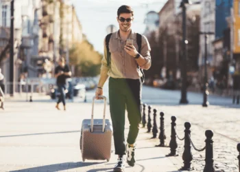 A young man wearing sunglasses pulls a suitcase through an unknown town. Using travel rewards cards can save you a ton on travel!