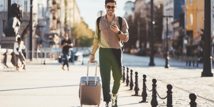A young man wearing sunglasses pulls a suitcase through an unknown town. Using travel rewards cards can save you a ton on travel!