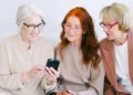 Three older women sit together, looking at a phone screen and talking about how to save money in retirement.