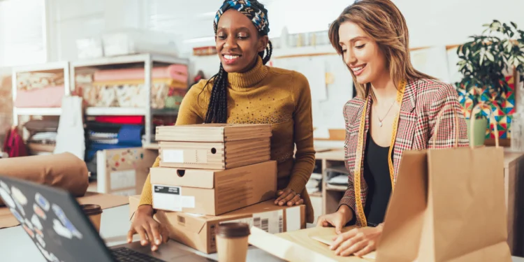 Two women pack up boxes at a counter. Selling at a craft show is one of the seasonal side hustles you can get extra cash from.