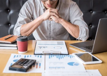 A man sits with his hands folded as he looks at his investments on his desk. Investing small is a good way to get started.