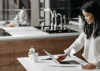 A woman with dark hair stands at a kitchen table, working on a laptop. Financial self-care is just as important as personal self-care.