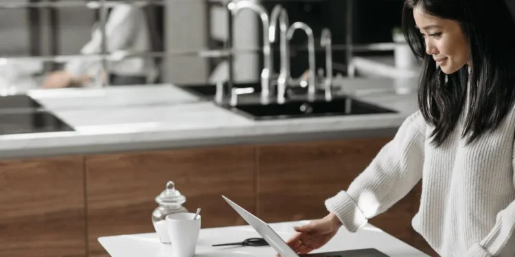 A woman with dark hair stands at a kitchen table, working on a laptop. Financial self-care is just as important as personal self-care.