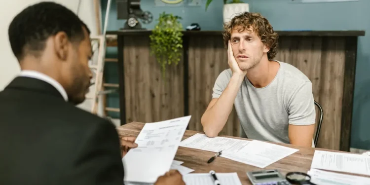 A man in a white shirt sits with his head in his hand across a desk from another man who is looking at paperwork. If you made a money mistake, get your plan ready ASAP to recover.