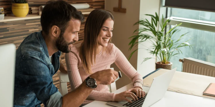 A man and woman smile as they look at a laptop. Buying and selling online can help put some money in your wallet, but you need to beware of scams.
