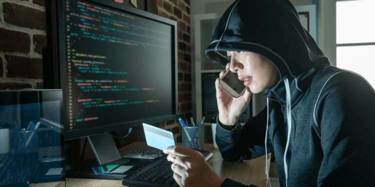 A person wearing a black hoodie sits in a dark room in front of a computer, looking at a bank card and talking on the phone. Holiday scams are in full force this time of year.