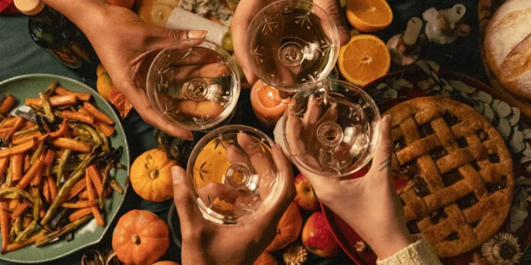 A group of hands toasts with glasses above a spread of food. A minimalist Thanksgiving can preserve your traditions while lowering stress, cost and expectations.