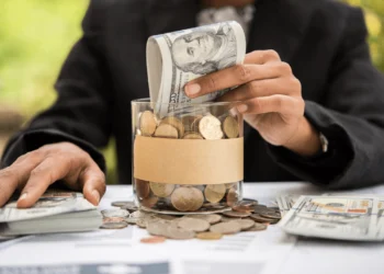 Hand stuffs dollar bills into glass jar filled with coins. Add $1 a week to your jar in the 52-Week Savings Challenge.