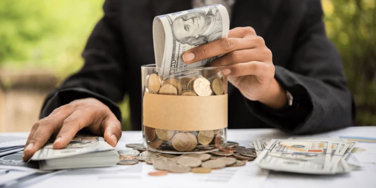 Hand stuffs dollar bills into glass jar filled with coins. Add $1 a week to your jar in the 52-Week Savings Challenge.