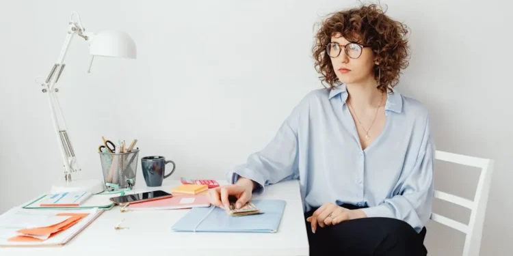 Woman wearing glasses holds a pile of money and sits at a table. Money dysmorphia is a distortion of your financial situation.
