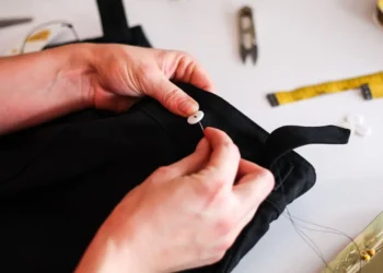 Hands stitch a button onto a pair of black pants. Repairing saves you money.