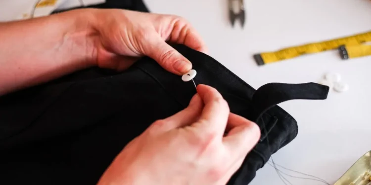 Hands stitch a button onto a pair of black pants. Repairing saves you money.