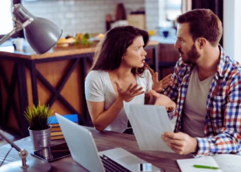 A man and woman look tense while looking at paperwork at a table. Living with an overspender can cause all kinds of drama.