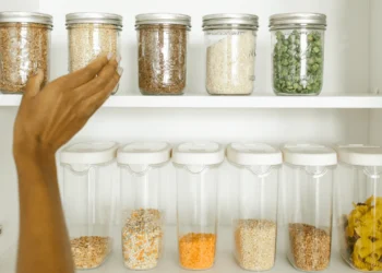 A hand places a jar back on a shelf in a tidy cupboard. Not having proper storage is one of the household things to stop wasting money on.
