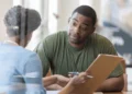 Man sits at a desk, looking at someone holding a clipboard.