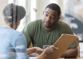 Man sits at a desk, looking at someone holding a clipboard.