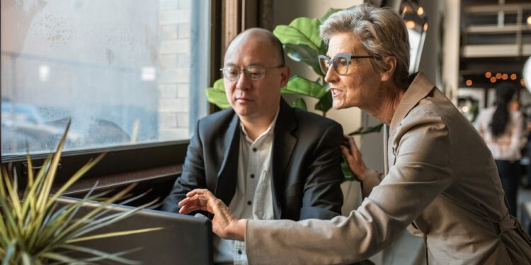 Woman stands over man at computer in a bright office.